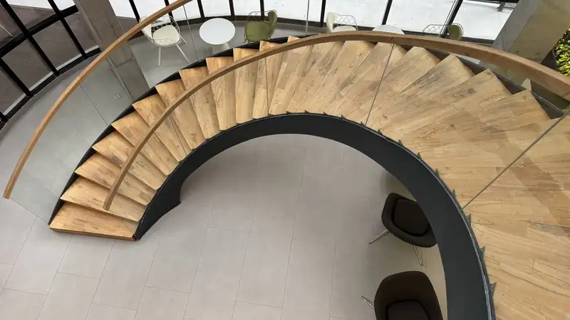 A curved wooden staircase with a glass railing, seen from above.