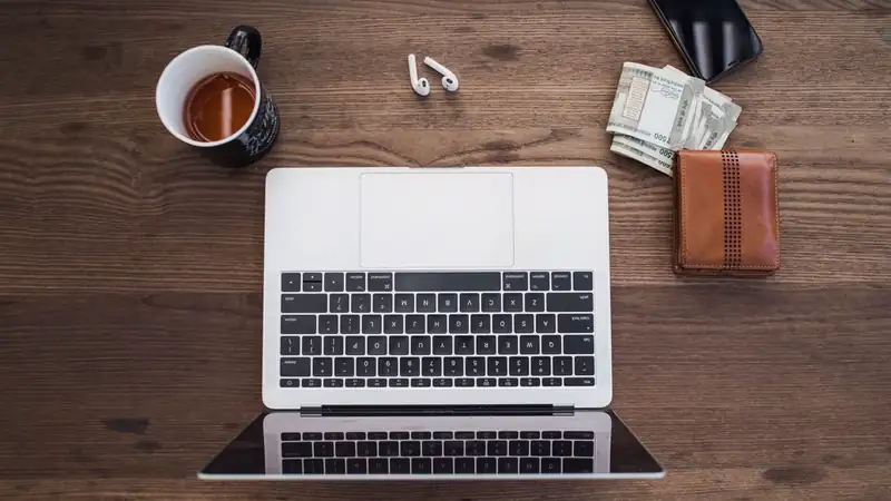 A laptop, a mug of tea, earbuds, folded dollar bills and a brown wallet on a wooden desk, seen from above.