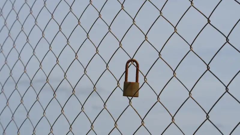 A small rusty padlock hanging on a chain-link fence.